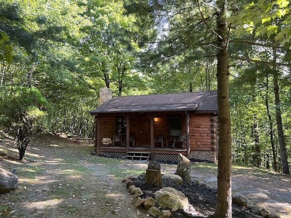 Une cabane en rondins dotée d’un porche, entourée d’arbres et bordée d’un chemin de gravier.