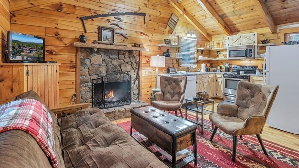 L’intérieur d’une cabane en rondins, avec une cheminée en pierre, des murs en bois, un coin cuisine et un salon comprenant un canapé et des fauteuils.