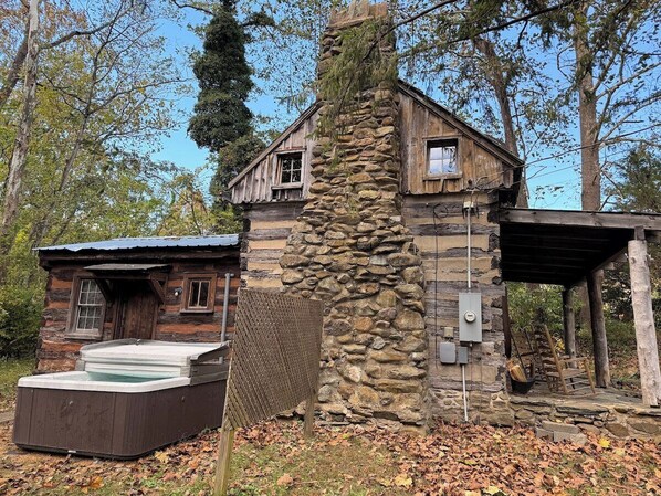 A rustic cabin with a stone chimney, a hot tub, and a covered porch.