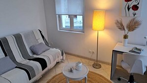 Ein Wohnzimmer mit einer Couch, einem kleinen runden Tisch mit Kerzen, einer Stehlampe, einem weißen Schreibtisch mit Stuhl und einem Fenster mit Ausblick.
