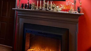 A fireplace with a lit fire, a mantel decorated with candles and flowers, and a red wall in the background. - Empty Arms Parlor - Rm. 208 (Stanardsville)