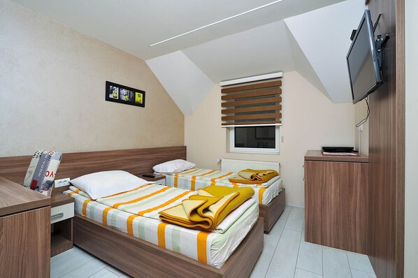 A compact hotel room with two beds, a small desk, a TV, and a window with blinds. - Vila Mina (Zlatibor)