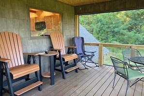 A covered outdoor seating area with Adirondack chairs and a table, a wooden deck, and a view of greenery.