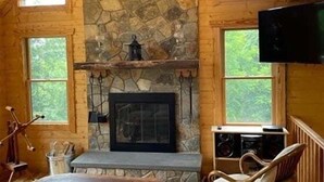 Ein Blockhaus-Interieur mit Kamin aus Stein, Holzwänden, einem an der Wand befestigten Fernseher und einem Fenster mit Blick auf Grünflächen.