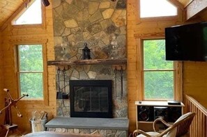 A log cabin interior with a stone fireplace, wooden walls, a television mounted on the wall, and a window overlooking greenery.