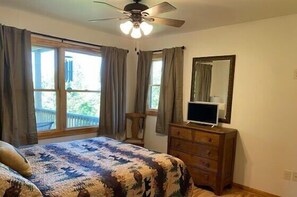 Un dormitorio con una cama, un aparador, un ventilador de techo, una ventana con cortinas y un espejo.