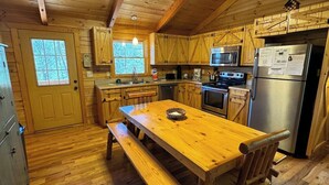 A wooden cabin interior with a wooden dining table, benches, a kitchen with wooden cabinets, a refrigerator, and a microwave. - Southern Comfort Cabin (Stanardsville)