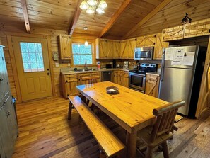 A wooden cabin interior with a wooden dining table, benches, a kitchen with wooden cabinets, a refrigerator, and a microwave.