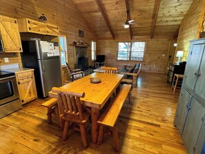 A wooden cabin interior with a dining table, benches, a refrigerator, a fireplace, and a television.
