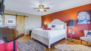 A bedroom with a bed, a ceiling fan, a desk, a chair, a red table, and a painting on the wall. - Hollywood Suite - Rm. 204 (Stanardsville)