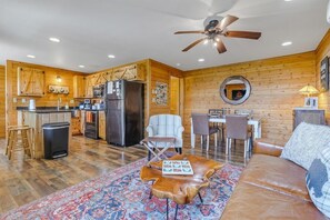 A spacious living area with a sofa, a coffee table, a dining table with chairs, a kitchen area with a refrigerator, and a ceiling fan.
