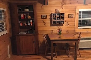 A wooden interior with a dining table, chairs, a small cabinet, and a window with blinds. - Tucked Away Mountain Cabin (Stanardsville)