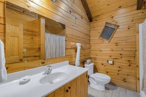 Baño de cabaña de madera con lavamanos, espejo, inodoro y una pequeña ventana.