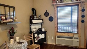 A kitchen with a stove, a dining table with chairs, a window with blinds, and a wall-mounted shelf with various items.