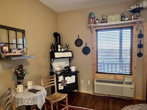 A kitchen with a stove, a dining table with chairs, a window with blinds, and a wall-mounted shelf with various items.