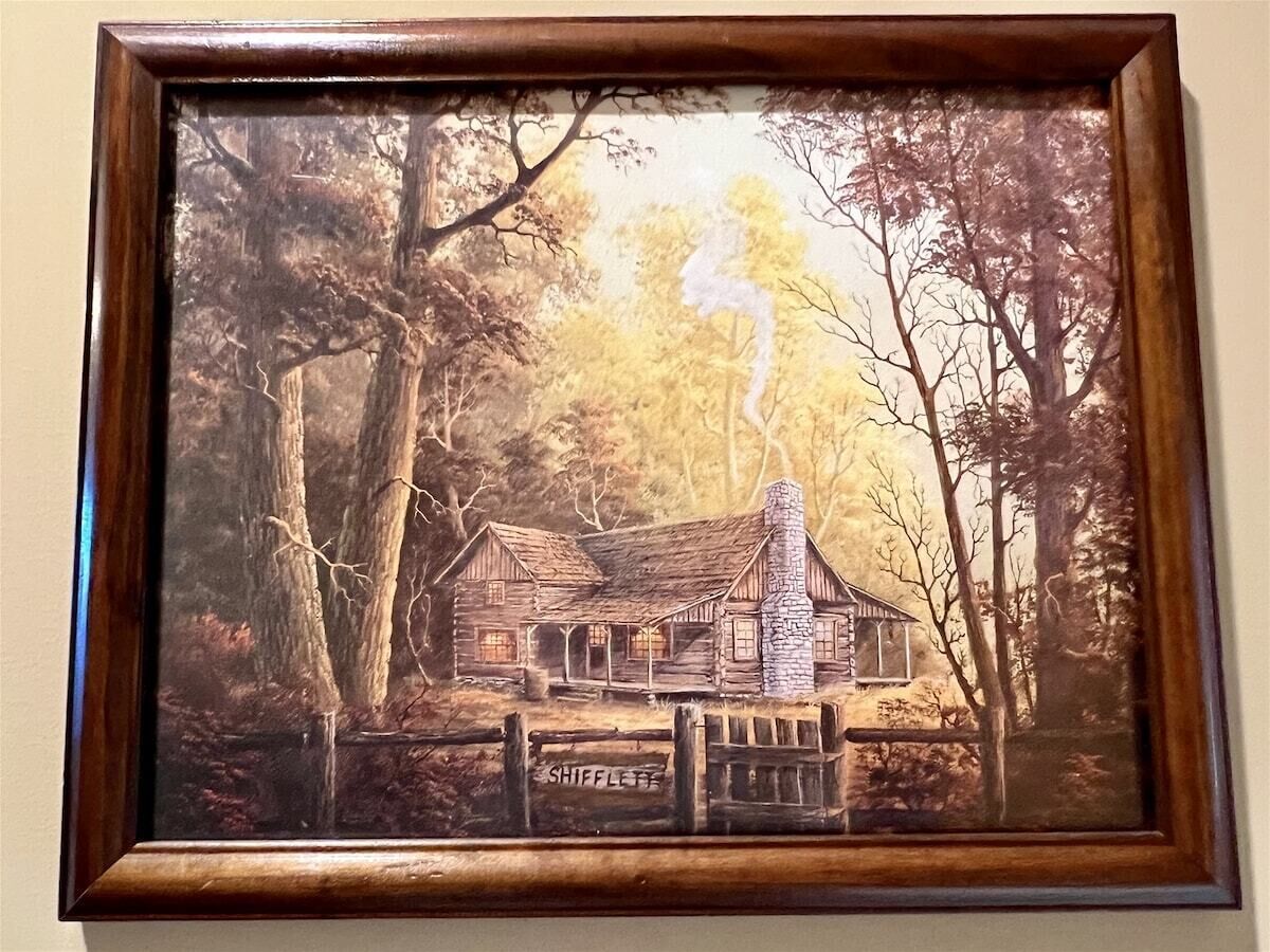 A framed painting of a rustic cabin with a chimney, surrounded by trees, and a sign that reads 'SHIFFLETT.'