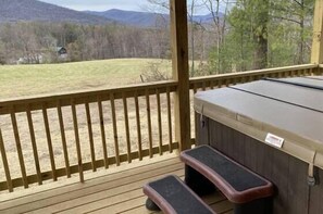 A deck with a hot tub, wooden railings, and a view of a grassy field and mountains.