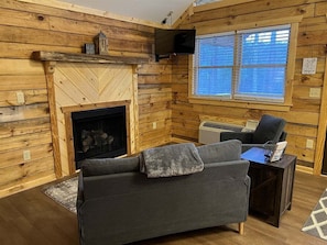 A cozy cabin interior with a fireplace, a sofa, a TV, and a window.