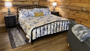 Een slaapkamer met een wand van houten panelen, een bed met een bedovertrek met patroon, twee nachtlampjes, een grijze bank en een klein vloerkleed.