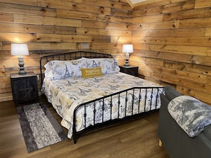 A bedroom with a wooden paneled wall, a bed with a patterned bedspread, two bedside lamps, a grey sofa, and a small rug.