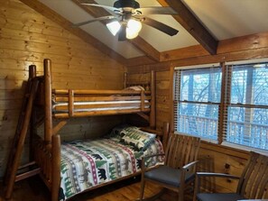 A wooden cabin room with a bunk bed, a single chair, a ceiling fan, and a window with blinds.