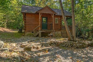 Une cabane rustique en rondins de bois, dotée d’un porche et entourée d’arbres et d’un mur de pierres.