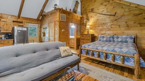 A log cabin interior with a sofa, a bed with a patterned blanket, wooden walls, a ceiling fan, and a kitchen area. - Cabin #5 - Love Nest (Stanardsville)
