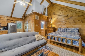 L’intérieur d’une cabane en rondins, avec un canapé, un lit recouvert d’une couverture à motifs, des murs en bois, un ventilateur de plafond et un coin cuisine.