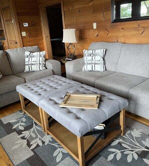 A cozy cabin interior with a grey sofa, a matching ottoman, and a wooden side table with a lamp.