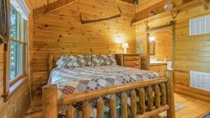 Interior de uma cabana de madeira com cama, janela, abajur e espelho.