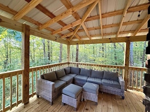 Un espacio de estar al aire libre techado con piso de madera, un sofá seccional grande y vista a un bosque.