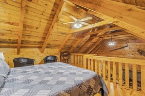 A wooden cabin with a high ceiling, a bed with a patterned bedspread, two chairs, a ceiling fan, and a railing along the upper wall.