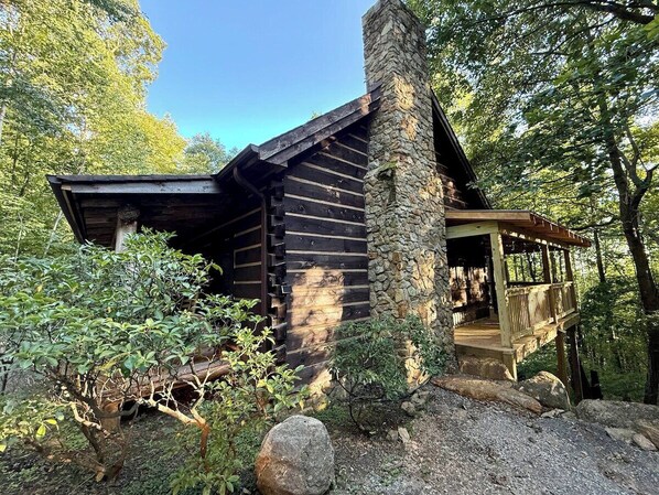Una cabaña de troncos con chimenea de piedra, un porche y un paisaje boscoso de fondo.