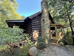 A log cabin with a stone chimney, a porch, and a forested backdrop. - Cabin #3 - Honey Pot (Stanardsville)