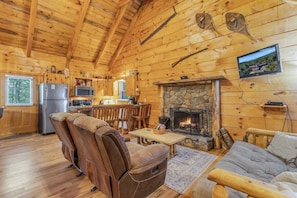 A log cabin interior with a stone fireplace, wooden furniture, a kitchen area, and a television mounted on the wall. - Cabin #3 - Honey Pot (Stanardsville)