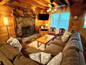 A cozy log cabin interior with a stone fireplace, a wooden ceiling, a television, and a sofa with cushions.