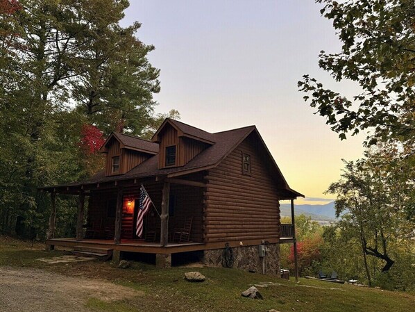 A rustic log cabin with a porch, a flag, and a view of the mountains.