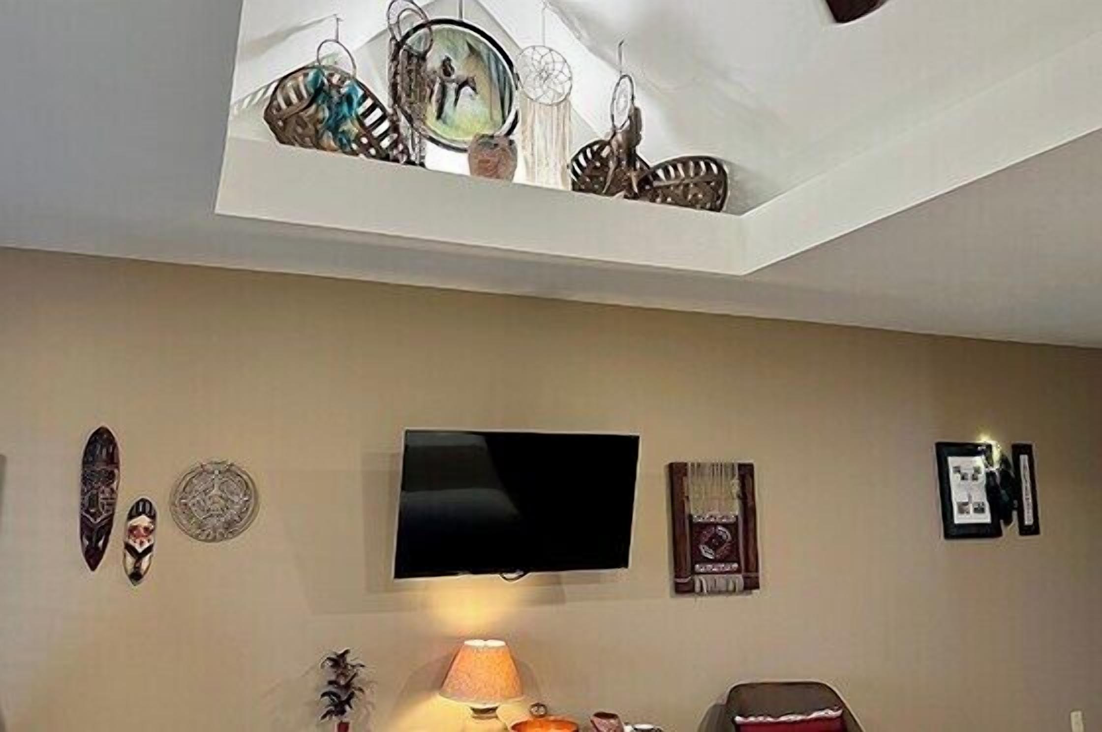 A room with a flat-screen TV mounted on the wall, decorative wall hangings, a lamp, and a small table.