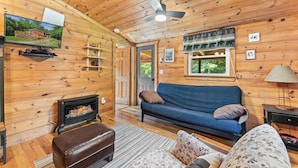 L’intérieur d’un chalet en bois, avec un canapé, un pouf, une cheminée et un ventilateur de plafond.