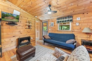 A wooden cabin interior with a sofa, ottoman, fireplace, and ceiling fan. - Mountain Escape Cottage (Stanardsville)