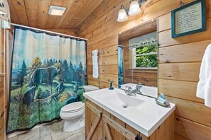 A wooden cabin bathroom with a forest-themed shower curtain, a white sink, and a mirror with a view of greenery outside. - Mountain Escape Cottage (Stanardsville)