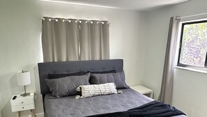 A bedroom with a bed, a nightstand, a lamp, a window with curtains, and a ceiling light. - Cozy apartment in Hollywood. Ideal Location Near Beach, Shops, and Highways (Hollywood)