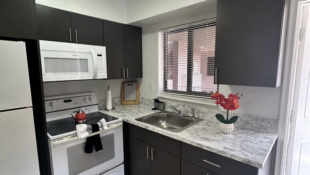 A modern kitchen with dark cabinets, a white refrigerator, a microwave, an oven, a sink, and a window with blinds. - Cozy apartment in Hollywood. Ideal Location Near Beach, Shops, and Highways (Hollywood)