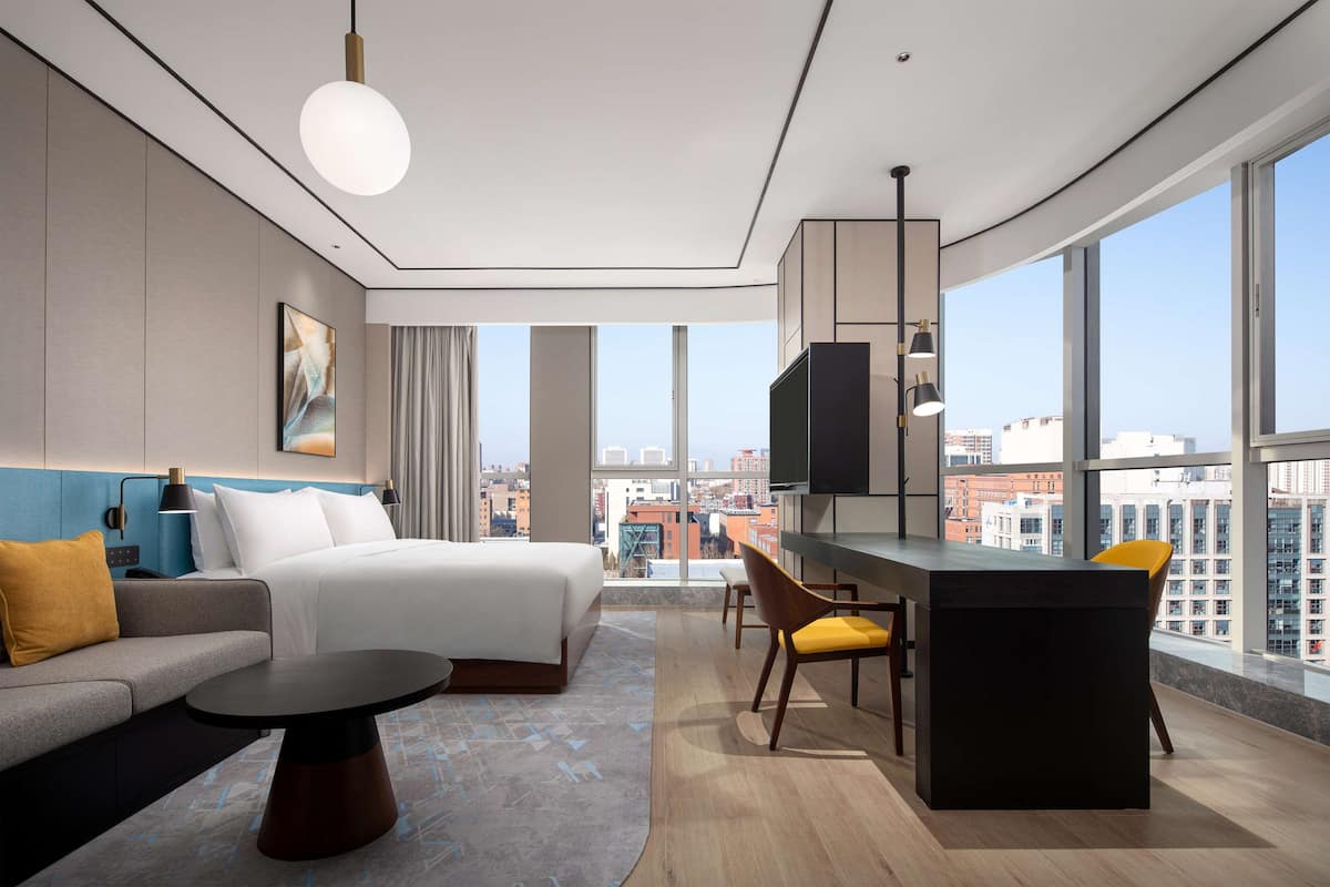 Una habitación de hotel moderna con cama, sofá, zona de comedor y amplios ventanales con vistas a la ciudad.