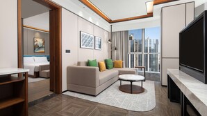 Una sala moderna con sofá, mesa de centro y televisor. Hay un comedor con mesa y sillas. La habitación tiene ventanas grandes con vista a la ciudad.