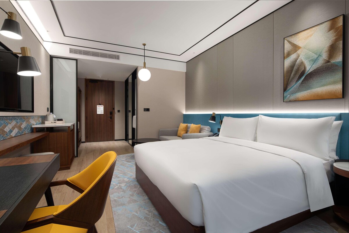 Une chambre d’hôtel moderne, dotée d’un grand lit, d’un bureau avec une chaise, d’un canapé et d’un grand tableau abstrait accroché au mur.