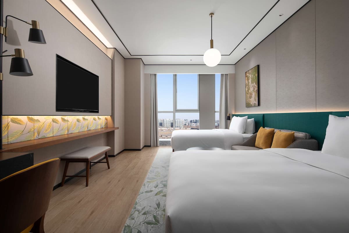 Habitación de hotel moderna con una cama grande, un escritorio con televisor de pantalla plana y vistas a la ciudad a través de amplias ventanas.