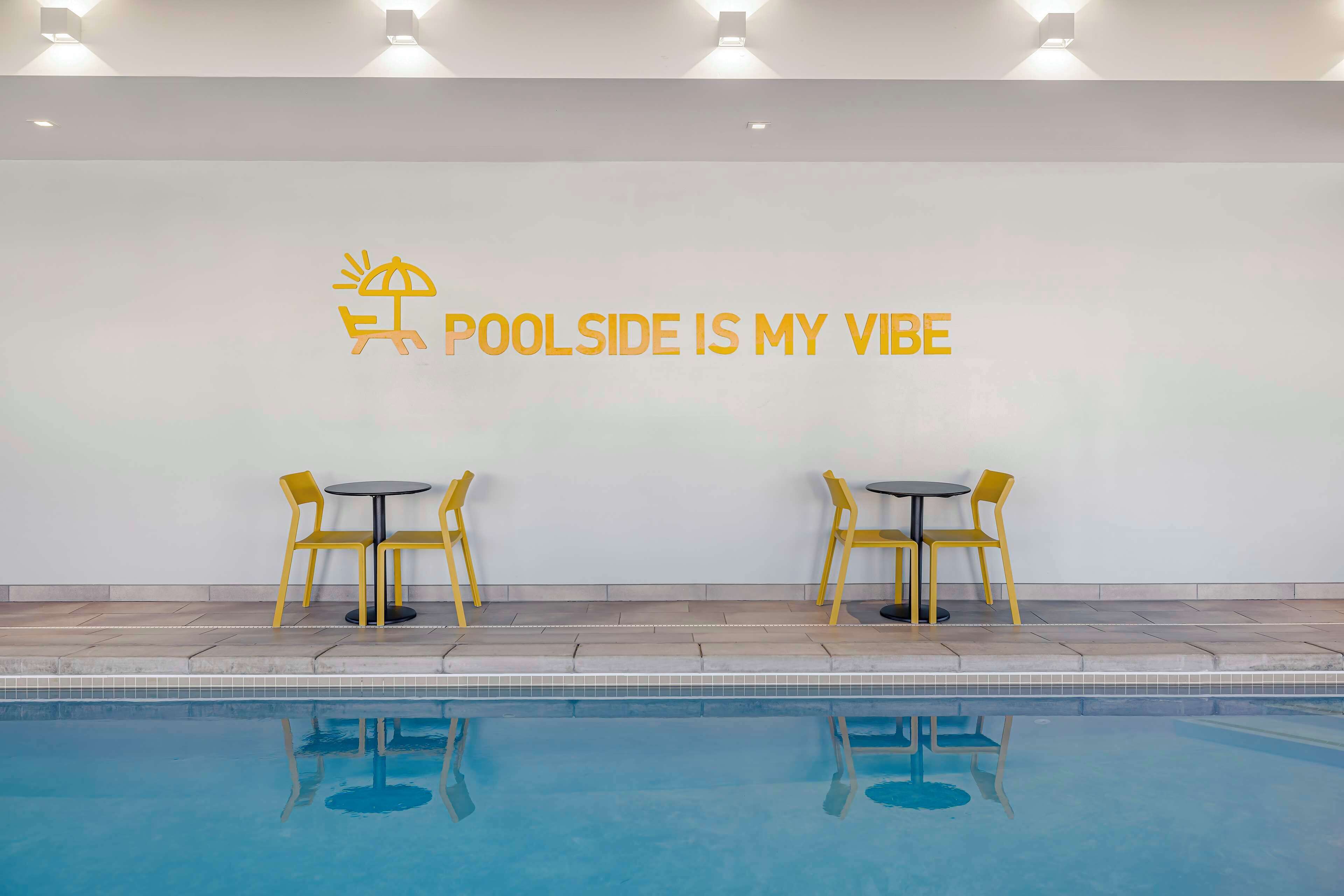 Ein Poolbereich mit einem Schild, auf dem 'POOLSIDE IS MY VIBE' steht, sowie zwei gelbe Stühle und ein Tisch.