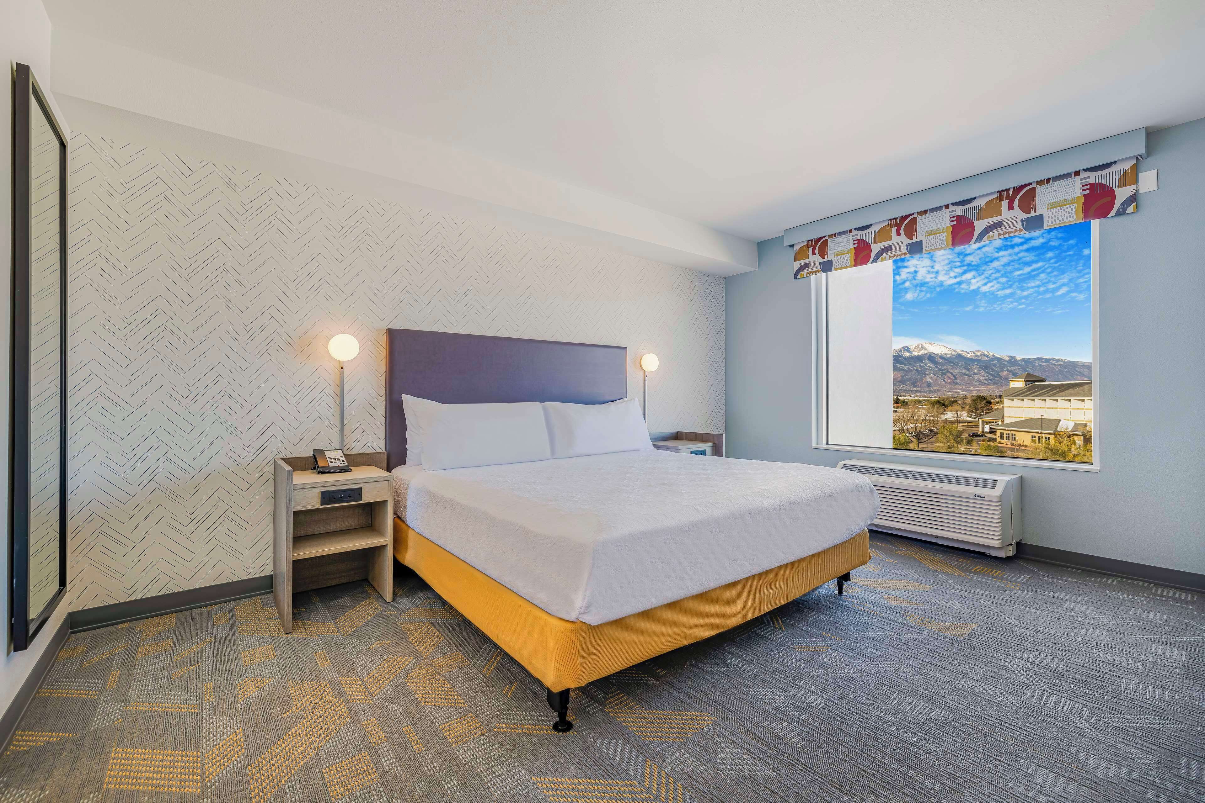 A hotel room with a bed, nightstands, a window with a mountain view, and patterned carpet.