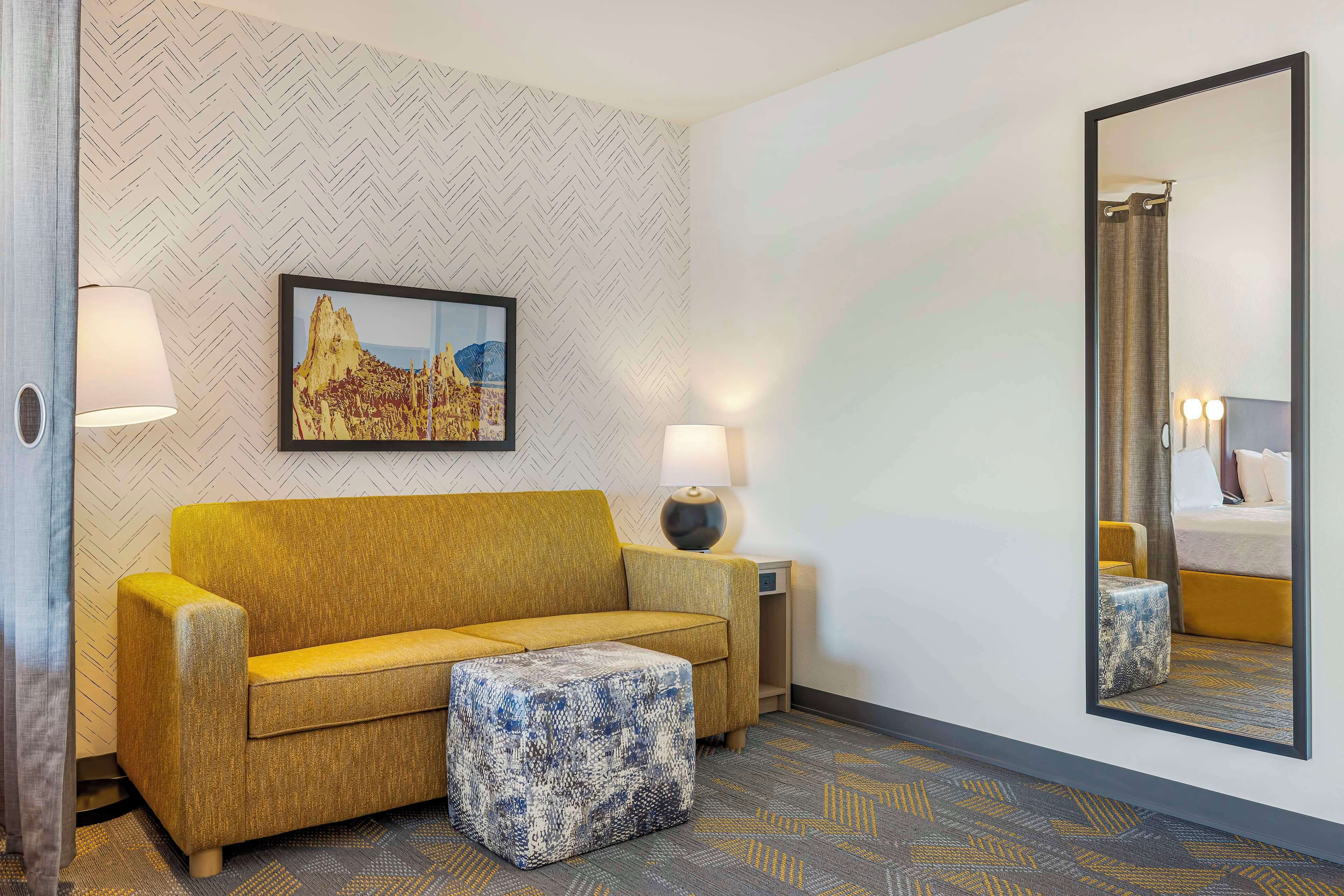 A hotel room with a sofa, ottoman, side table, lamp, and a large mirror.